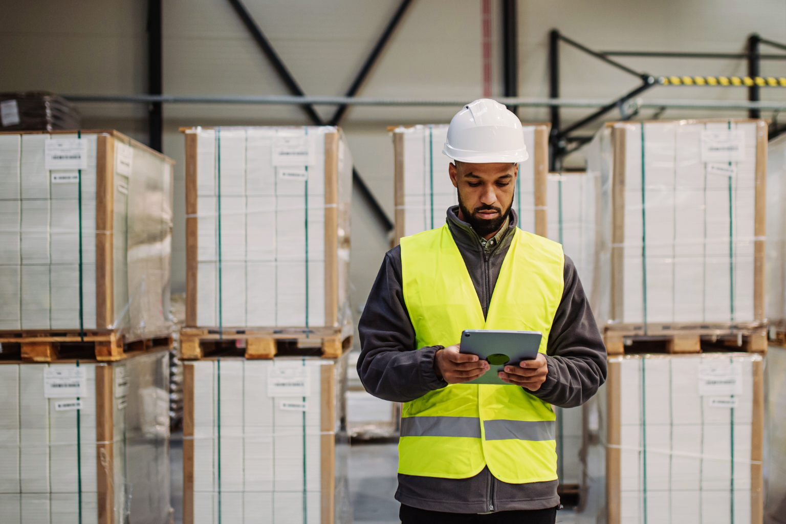 A man in a yellow safety vest and white hard hat is using a tablet at the UPS facility warehouse. Pallets of stacked boxes are visible in the background as he checks updates on a potential delivery delay.