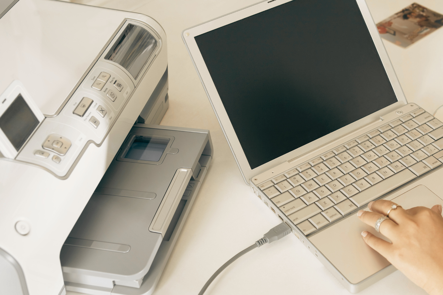 A person using a white laptop, possibly to access FedEx Ship Manager, is connected to a printer. The laptop screen is blank, with their hand on the keyboard. The printer, which may reprint a label or a FedEx label, displays several buttons and a small display screen on the white surface.