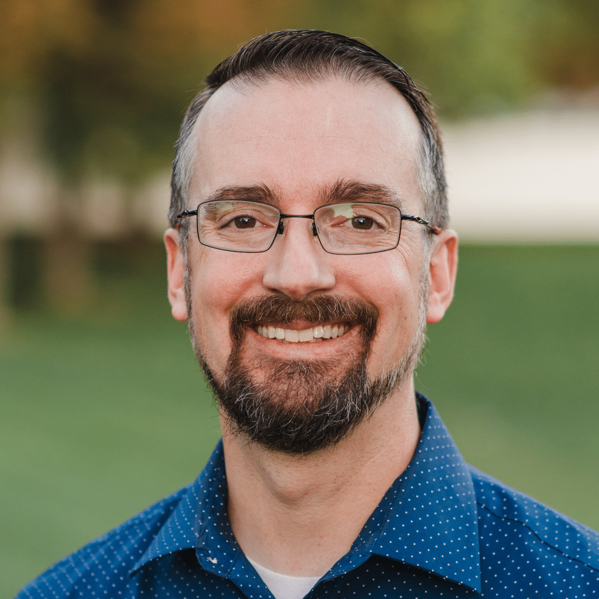A man with short brown hair, glasses, and a trimmed beard smiles at the camera. He is wearing a blue button up shirt with small white dots. The background is blurred greenery.