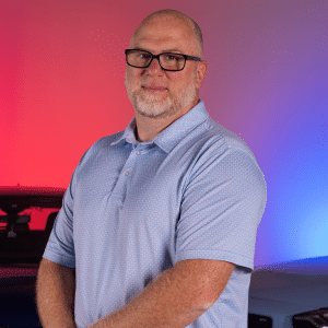 A man with short hair, glasses, and a beard wearing a light blue polo shirt stands in front of a blurred background with red and blue lighting, representing ECS Tuning’s expertise in high volume shipping.
