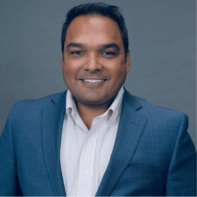 A man with dark hair wearing a blue suit jacket and a light colored checkered shirt is smiling at the camera against a plain gray background.
