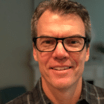 A middle aged man with short brown hair, wearing black rimmed glasses and a dark plaid shirt, smiles at the camera. The background is softly blurred.