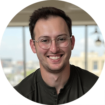 A young man with short dark hair and glasses smiles at the camera. He is wearing a dark shirt and is indoors, with large windows and a blurred cityscape in the background.