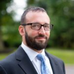 A man with short brown hair, a trimmed beard, and glasses is wearing a dark suit, white shirt, and blue patterned tie, standing outdoors with greenery in the blurred background.