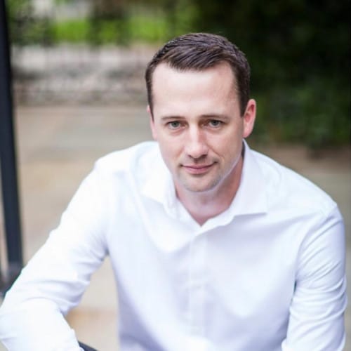 A man with short brown hair, wearing a white button up shirt, sits outdoors on a stone surface with greenery in the background, looking up at the camera with a slight smile—perhaps pondering ShipScience and carrier reimbursements.